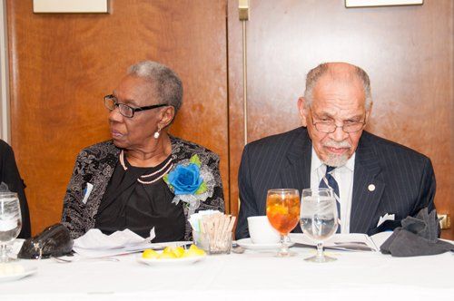 Two people seated at a table. A woman in a black dress looks left, while a man in a suit looks down.