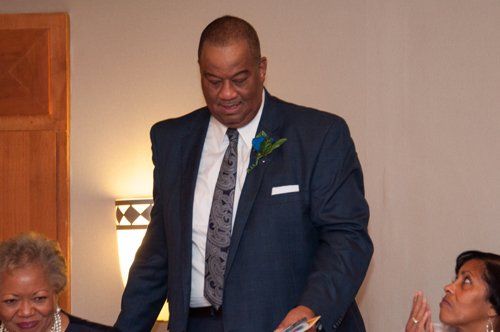 Man in a blue suit at an event, holding a card, looking down. Others are seated nearby.