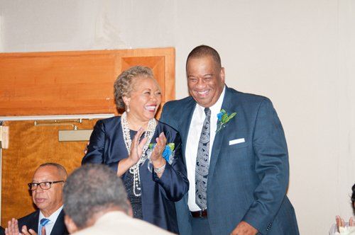 Woman in navy jacket and man in suit smile, clapping in a room; another man sits in background.