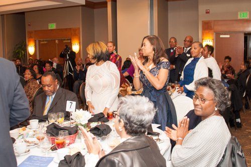 People at a formal event, clapping. Elegant attire, tables set with flowers and food, indoors.