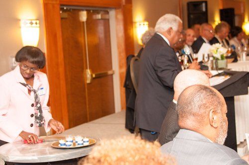 Woman arranging pastries on a table at a formal event, other attendees behind a table.
