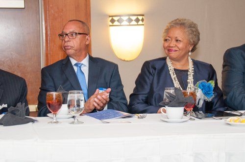 Man in suit clapping, woman in suit smiling at table setting, white tablecloth.