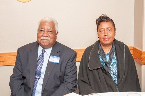 An older man and woman seated indoors, smiling. Man in suit, woman in dark coat.