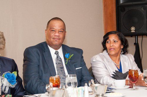 Man in blue suit, woman in white blazer at a formal table. Both smile, likely at an event.