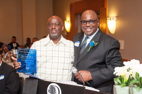 Two men at a podium; one holding an award, other in a suit with a flower, inside a banquet hall.