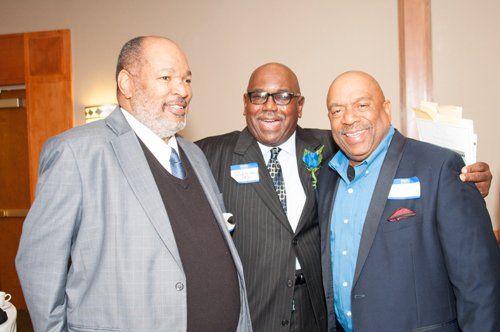 Three men smiling, arm-in-arm. Two wear suits; one a blue blazer and button-down shirt. Beige room, tags on lapels.