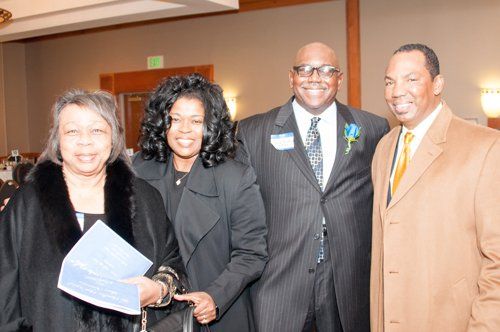 Four formally dressed people smiling indoors, posing closely together.