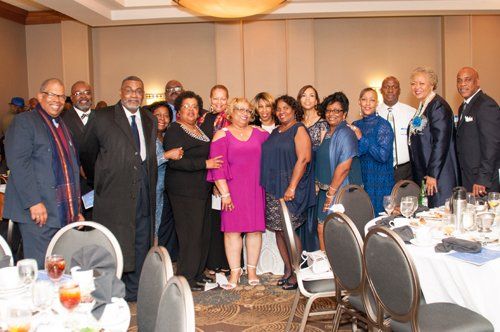 Group of people at a formal event, smiling, posing near tables, wearing suits and dresses.