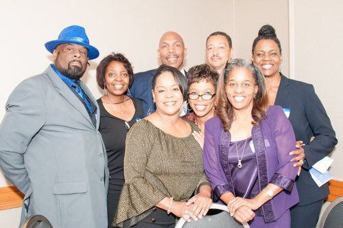 Group of people posing for a photo at an event, smiles, variety of outfits, indoors, neutral background.