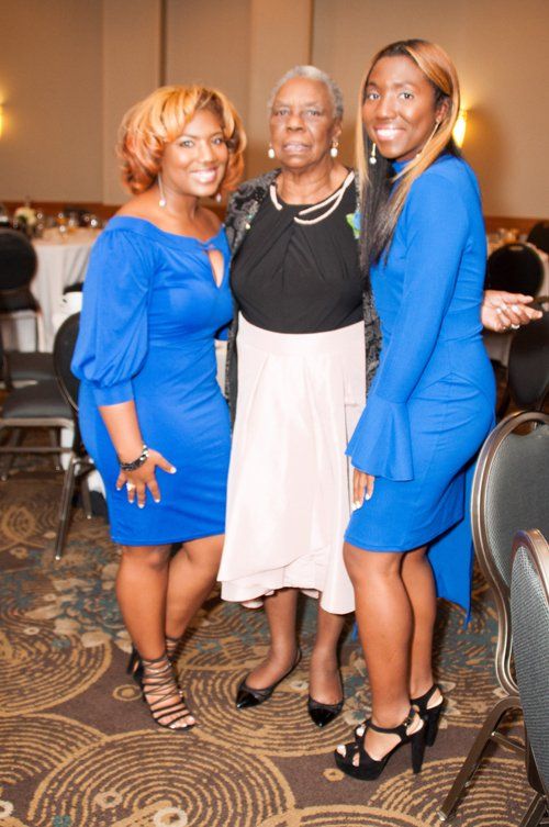 Three people posing for a photo at an event; two women in blue dresses flank an older woman in a black and beige outfit.