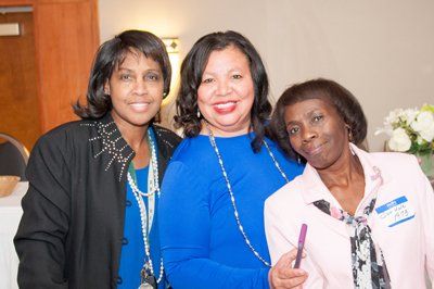 Three women smiling together indoors, one in blue, one in pink, and one in a black jacket.