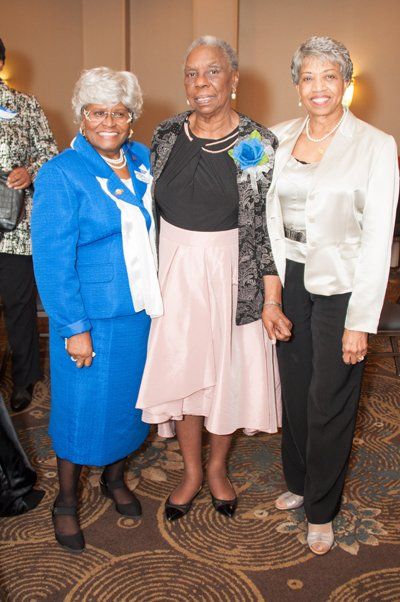 Three women in formal attire, smiling, indoors. One in blue suit, one in black and pink, one in white jacket and black pants.