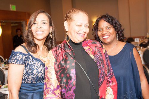 Three women smiling at a gala. One wears a floral jacket, others in blue dresses.