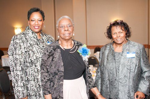 Three women in formal attire at an event, smiling. One wears a blue flower. Indoors, neutral background.
