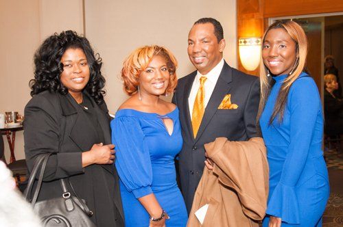 Four people posing for photo indoors. Woman in black blazer, blue dress, man in suit, and woman in blue dress.