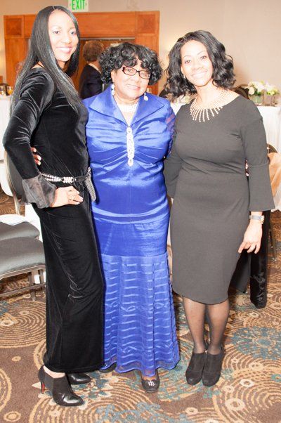 Three women pose together indoors. One in blue, the others in black.
