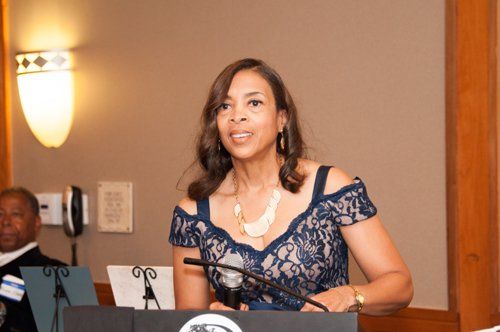 Woman speaking at a podium, wearing a blue lace dress, with a microphone. Another person is seated to the left.