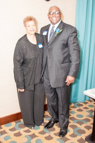 Woman and man in formal attire pose together indoors. Man in pinstripe suit, woman in black pantsuit.