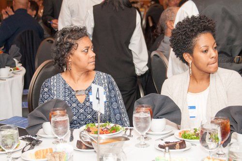 Two women seated at a formal dinner table, looking to the side. Food and drinks are on the table.