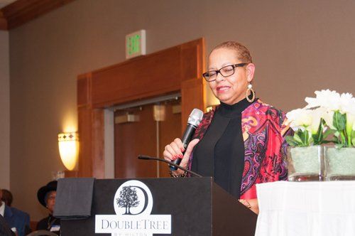 Woman speaking at a DoubleTree podium, holding a microphone. She wears glasses and a patterned jacket. Interior setting.