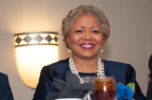 Smiling person with pearl necklace at a formal event, lit by a wall sconce.