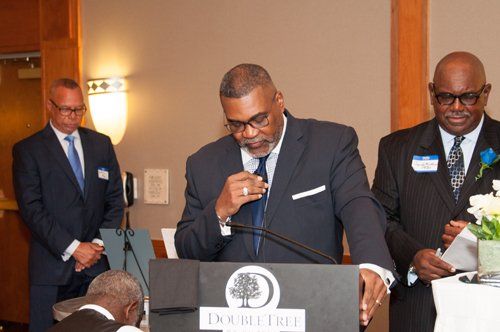 Three men at a podium; one speaks, others listen. The setting is indoors, likely a formal event.