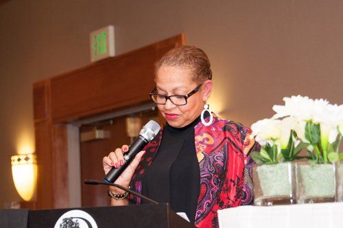 Woman speaking at a podium with microphone. She is wearing glasses and a colorful jacket, speaking in a room.
