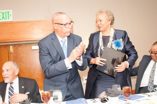 Man in blue suit applauds woman holding a folder, standing at a table. Others seated.