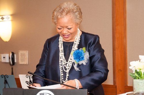 Woman in navy jacket speaks at podium, wearing pearls and a blue flower; interior setting.