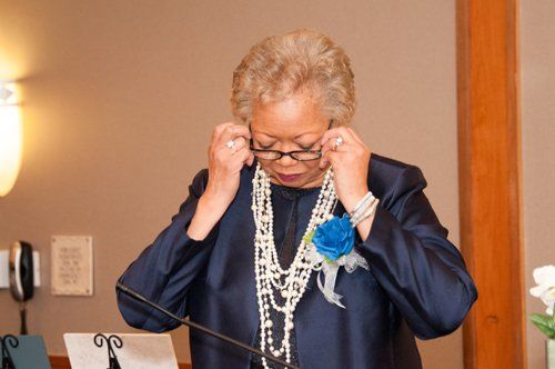 Woman in blue suit adjusting glasses, standing near a lectern, pearls around her neck.