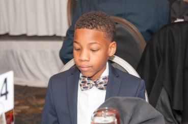 Boy in a navy suit and patterned bow tie sits at a table, looking down.