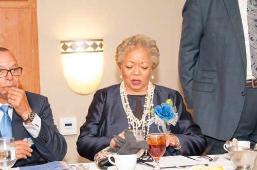 Woman in navy blazer at a table, holding object, with other people and a flower decoration.