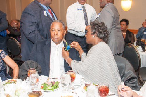 Woman pinning a blue ribbon on a man's lapel at a seated dinner. Other people are in the background.