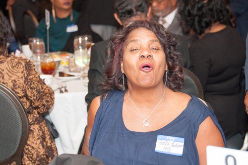 Woman at a table with her mouth open; wearing a blue top, a necklace, and a name tag that reads 