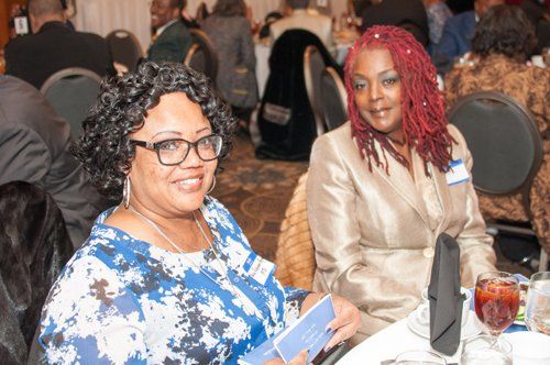 Two women at a formal dinner, one in a blue floral top, the other in a gold jacket. Both smile at the camera.