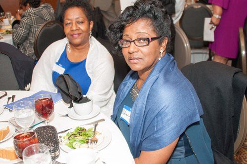Two women seated at a table at an event. The woman in the foreground wears a blue shawl and glasses.