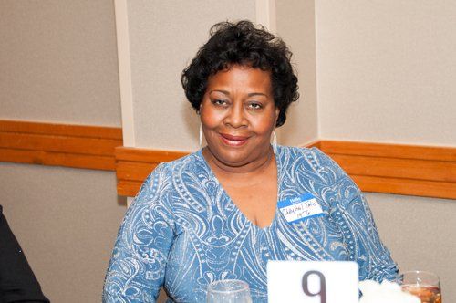 Woman with short, dark hair, wearing a blue patterned top, smiles at a table.