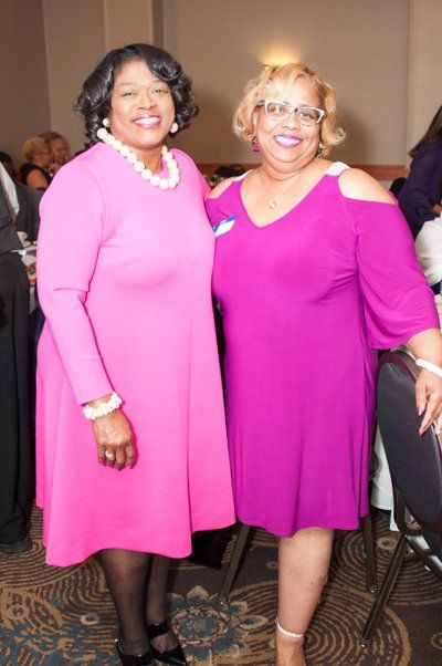 Two women pose together; one in a pink dress, the other in a magenta dress. They're at an event, smiling.