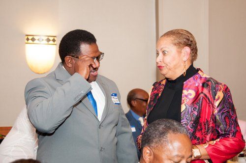Man in suit touching face, talking to woman in patterned jacket. Indoors, other people in background.