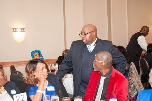 People at a formal event; man in suit stands, others seated at tables. Dim lighting.