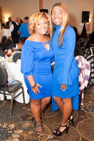 Two women in blue dresses pose at an event. They are smiling. The background has people and tables.