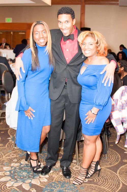 Three people posing for a photo. A man in a suit and two women in blue dresses smiling, indoors.