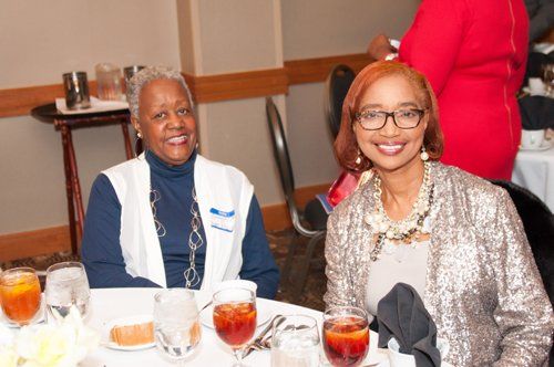 Two women seated at a table, smiling. One in blue vest, the other in a sequined jacket, with drinks and food.