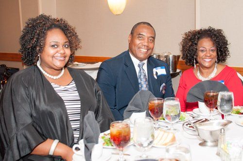 Three people seated at a table, smiling. Man in suit between two women. Drinks and food visible.