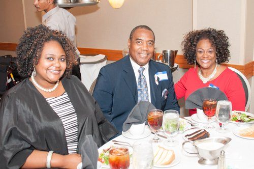 Three people smiling at a table set for dinner. The man in a suit, two women with curly hair.
