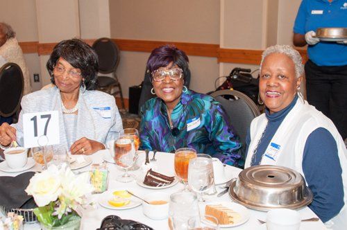 Three women smiling at a table set for a meal; table number 17, drinks, food, server in background.
