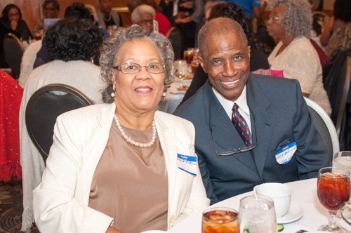Couple smiling at a formal event, seated at a table with drinks and other attendees in the background.