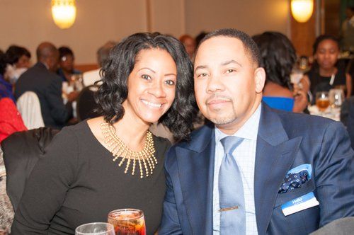 Woman in black dress and man in blue suit smile at a formal event, restaurant setting.