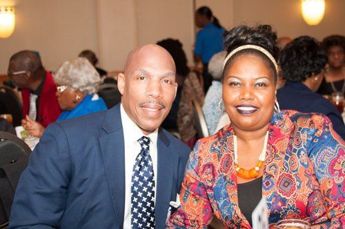 Couple smiles for the camera at a banquet. The man wears a blue suit; woman a patterned jacket. Others dine in background.
