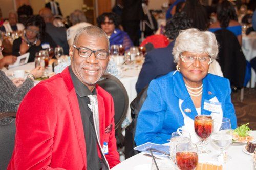 Man in red blazer and woman in blue jacket seated at a table, smiling, at a formal event.